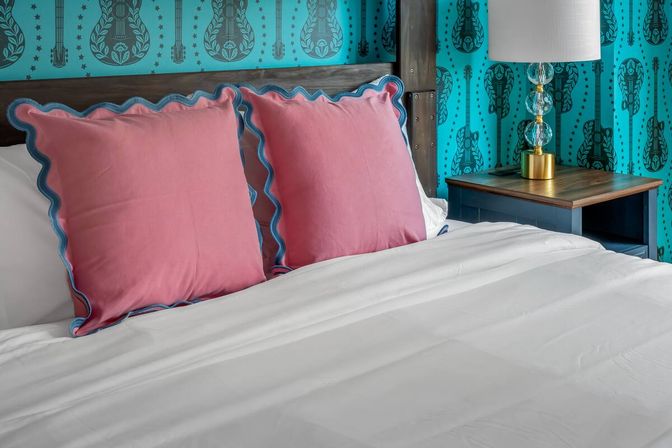 Cozy bedroom with crisp white linens, two pink scalloped-edge pillows with blue trim, rustic wood headboard, turquoise guitar-print wallpaper, and a wooden nightstand with a glass lamp.
