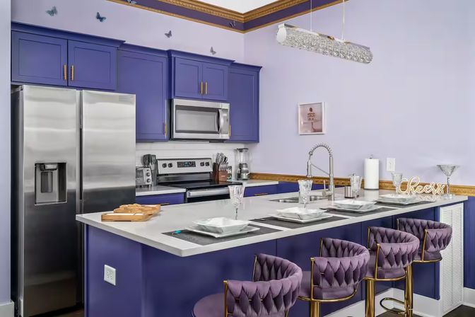 Modern purple kitchen with white quartz island, stainless steel appliances, tufted velvet purple bar stools with gold frames and place settings at the breakfast bar