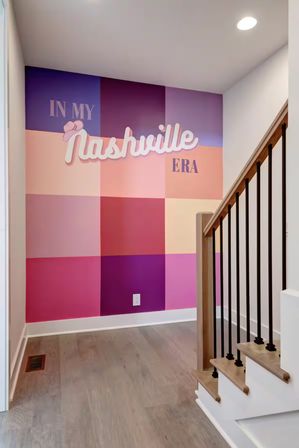Colorful pink and purple checkerboard wall mural reading 'In My Nashville Era' at a bright stair landing with wood-and-black metal railing and light hardwood floor.