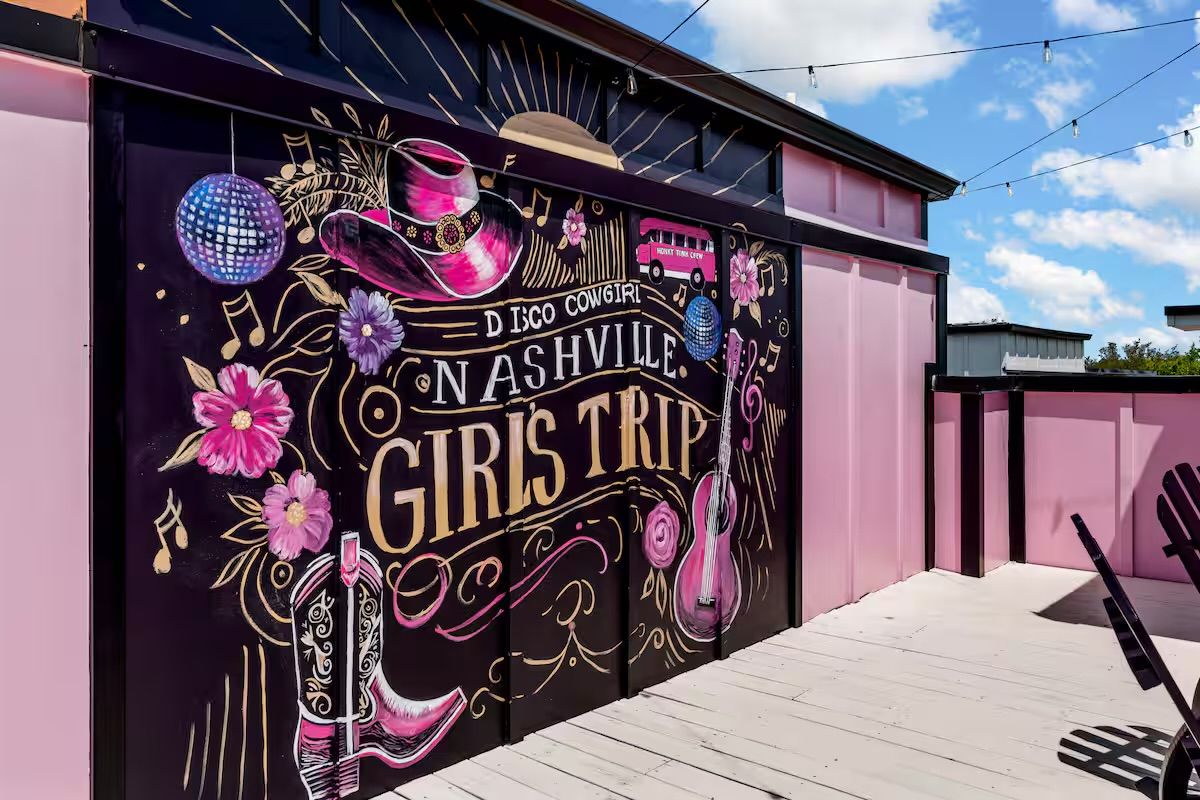 Vibrant Nashville rooftop mural reading “Girls Trip” in disco-cowgirl style — pink cowboy hat and boots, acoustic guitar, disco ball and floral accents on black and pink panels beneath string lights and blue sky.