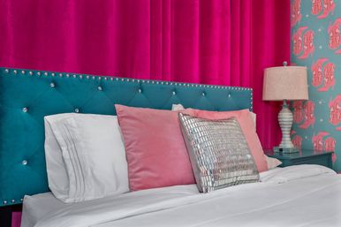 Stylish glam bedroom with a teal tufted headboard studded with crystals, hot-pink velvet backdrop, white bedding, blush pillows and a mirrored metallic throw pillow on a teal nightstand beside patterned pink-and-teal wallpaper and a lamp.