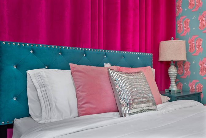 Stylish glam bedroom with a teal tufted headboard studded with crystals, hot-pink velvet backdrop, white bedding, blush pillows and a mirrored metallic throw pillow on a teal nightstand beside patterned pink-and-teal wallpaper and a lamp.