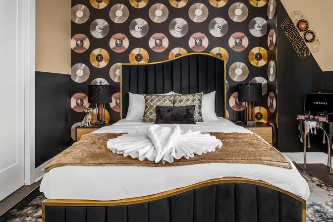 Music-themed boutique hotel bedroom with king bed, black velvet and gold headboard, gold-and-silver vinyl record wallpaper, swan towel on a brown throw, bedside lamps and retro decor.