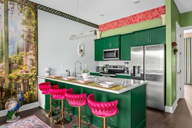 Bright open kitchen with emerald green cabinets, white island with four hot-pink velvet bar stools, stainless steel appliances and a scenic garden mural wall