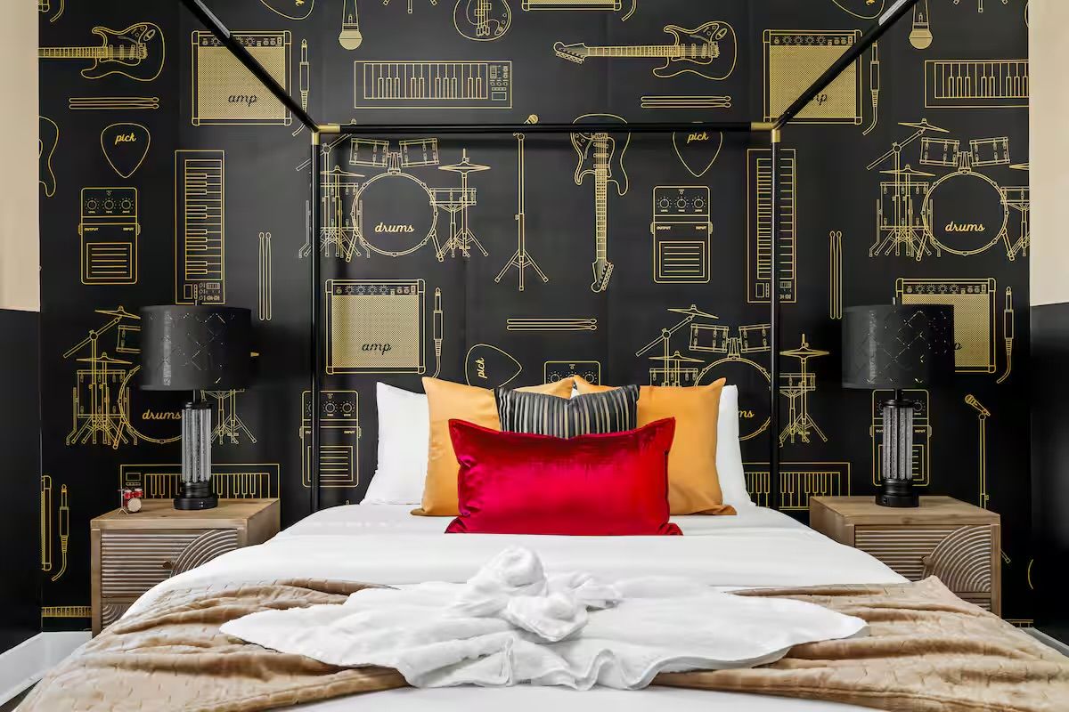 Contemporary music-themed boutique hotel bedroom with black-and-gold instrument wallpaper, four-poster bed topped with a bold red velvet pillow and golden cushions, wooden nightstands with black lamps, and a plush beige throw.
