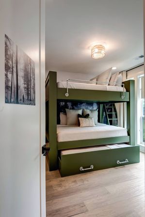Cozy contemporary bedroom featuring an olive-green built-in bunk bed with ladder and pull-out trundle, white bedding and accent pillows, forest wall art, hardwood floors, large window with curtains, and a woven ceiling light.