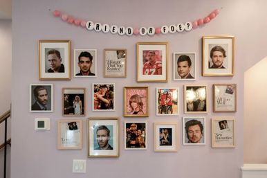 Pastel-pink home hallway gallery wall with a tidy grid of framed portraits, headshots and magazine-cover photos beneath a pink bead banner reading 'FRIEND OR FOE?'