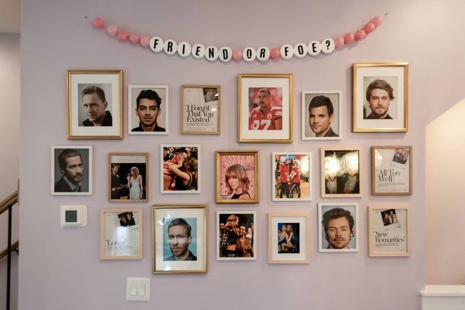 Pastel-pink home hallway gallery wall with a tidy grid of framed portraits, headshots and magazine-cover photos beneath a pink bead banner reading 'FRIEND OR FOE?'
