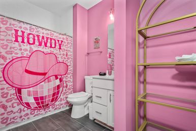 Playful bright pink bathroom with a 'HOWDY' leopard-print shower curtain showing a cowboy hat over a disco ball, white toilet and vanity, gray floor tiles, and gold metal shelving.