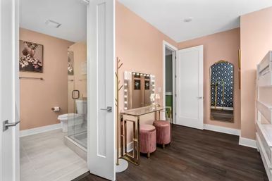 Modern glam pink dressing area with lighted vanity mirror, gold-framed glass desk, two pink velvet stools, full-length decorative mirror, and adjacent glass shower bathroom.