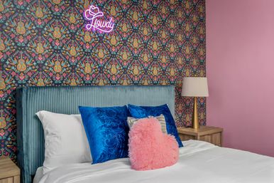 Playful bedroom scene with teal velvet headboard, colorful floral wallpaper and neon 'Howdy' sign, white bedding topped with blue satin pillows and a pink fuzzy heart cushion, wood nightstand and gold lamp