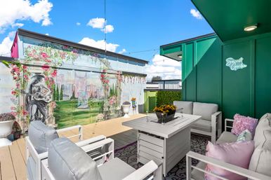 Sunny urban rooftop patio with white cushioned seating and fire-table, green accent wall with 'Secret Garden' sign, floral mural backdrop and string lights.