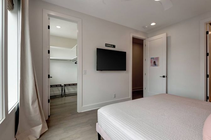 Bright modern bedroom with hardwood floors, pale pink quilted bed, wall-mounted TV, open walk-in closet and sunlit window with drapes