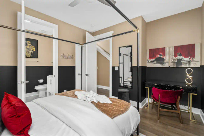 Modern bedroom with black-and-tan two-tone walls, four-poster bed dressed in white linens with red accent pillows and tan throw, hardwood floors, red velvet desk chair at a black console, full-length mirror, and open door to an en-suite bathroom.