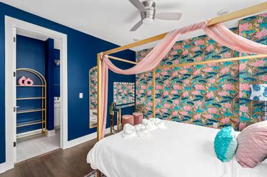 Colorful modern bedroom with gold four-poster canopy bed draped in pink fabric, tropical floral wallpaper, navy blue accent wall, white bedding with teal and pink pillows, vanity with round mirror and pink stools, gold shelving, hardwood floor and silver ceiling fan