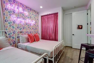 Pink-themed bedroom with two white metal twin beds, geometric pastel wallpaper, neon script sign saying And by the way I'm going out tonight, sparkly pink curtain and coral throw pillows on hardwood floor.
