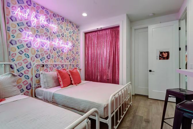 Pink-themed bedroom with two white metal twin beds, geometric pastel wallpaper, neon script sign saying And by the way I'm going out tonight, sparkly pink curtain and coral throw pillows on hardwood floor.