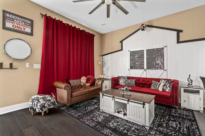 Music-themed modern farmhouse living room with red velvet sofa, brown leather chesterfield, white barn-style accent wall with sliding doors, bold red curtains, cowhide ottoman, rustic coffee table and ceiling fan.