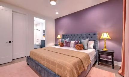 Cozy contemporary bedroom with tufted gray headboard against a purple accent wall, beige quilted comforter, velvet and knot throw pillows, dark nightstands with brass lamps, and an open doorway to an ensuite bathroom.
