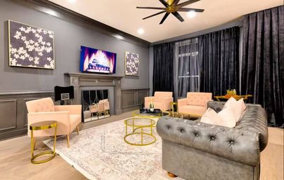 Modern living room with gray paneled walls, tufted leather sofa, blush armchairs, gold accent tables, wall-mounted TV above fireplace and dark velvet curtains.