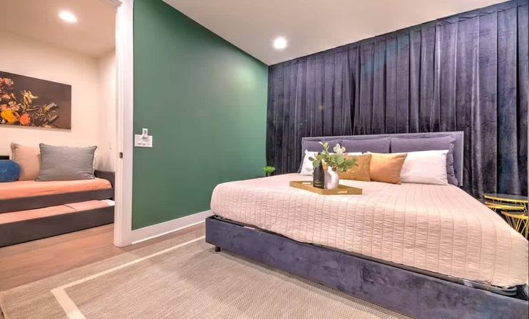 Cozy modern bedroom interior with velvet gray upholstered bed and pleated dark curtains, blush quilted bedding, green accent wall, small seating nook with sofa and floral art, beige area rug and decorative tray with vase.