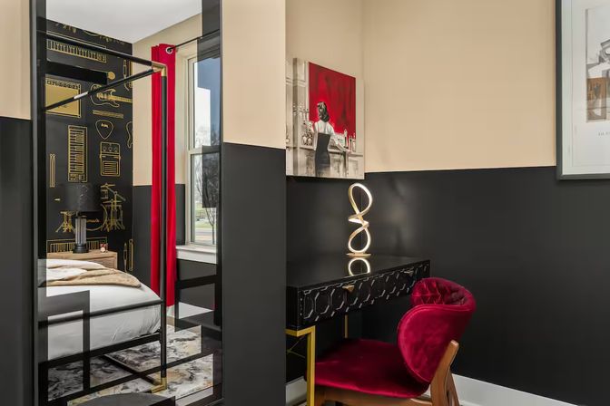 Stylish urban bedroom nook with black-and-cream walls, red velvet chair, black geometric desk topped by a looping LED lamp, wall art and tall mirror reflecting a bed with gold-accent musical wallpaper and red curtains.