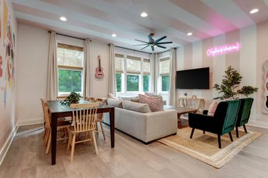 Bright contemporary open-plan living-dining room with bay windows, light-gray sectional sofa, green velvet armchairs, wooden dining table, wall-mounted TV, pink neon sign and hanging acoustic guitar.