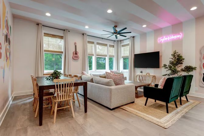 Bright contemporary open-plan living-dining room with bay windows, light-gray sectional sofa, green velvet armchairs, wooden dining table, wall-mounted TV, pink neon sign and hanging acoustic guitar.