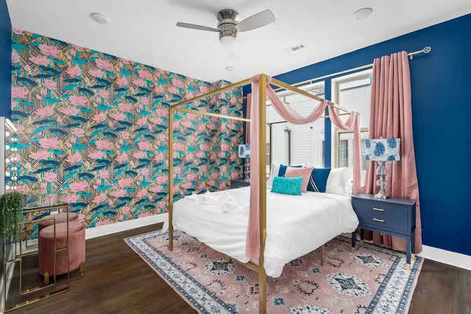 Vibrant modern bedroom with a gold four-poster canopy bed draped in pink fabric, tropical pink-and-teal floral wallpaper, navy accent wall, pink curtains, patterned pink area rug, blue nightstand with lamp, and dark hardwood floors.