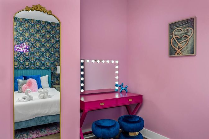 Boutique-style pink dressing nook with a hot-pink vanity and illuminated Hollywood mirror, two blue velvet stools, and a gold-trim full-length mirror reflecting patterned wallpaper and a white bed with plush pillows and towel swans.