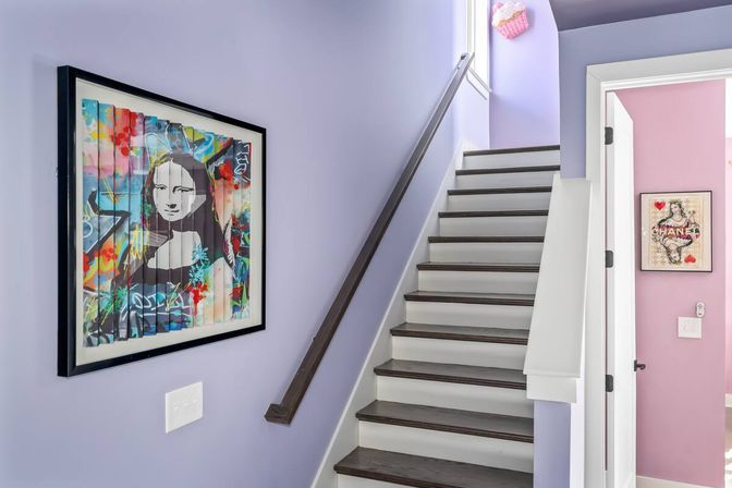 Lavender stairwell in a modern home with dark wood treads and white risers, a simple dark handrail, a colorful framed pop-art portrait on the wall, and a peek into a pink bedroom with additional artwork.