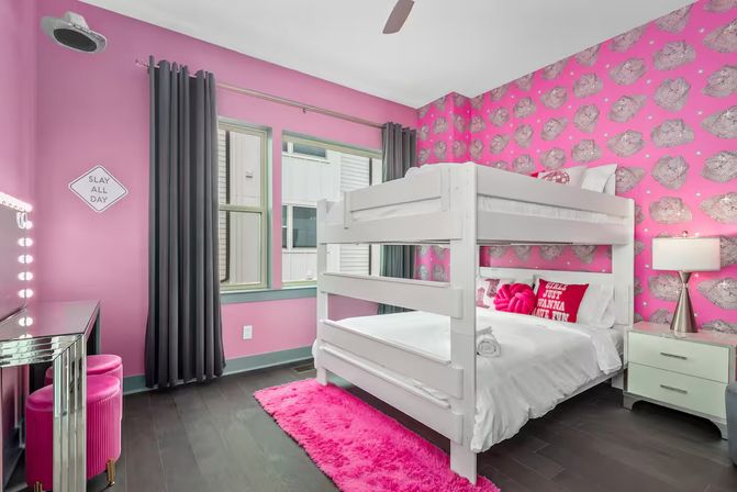 Playful bright pink kids' bedroom with white bunk bed, pink cowboy-hat wallpaper, hot pink shag rug, lighted vanity with pink stools, gray curtains and dark hardwood floor.