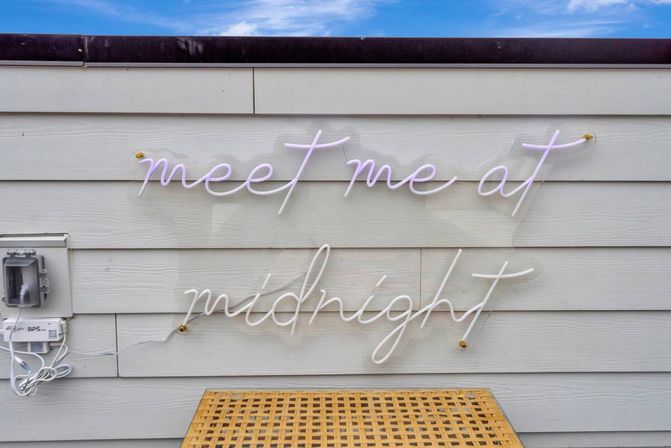 Playful cursive neon sign reading 'meet me at midnight' mounted on light gray horizontal siding above a woven rattan table, with an electrical box and cords to the left and a sliver of blue sky above.
