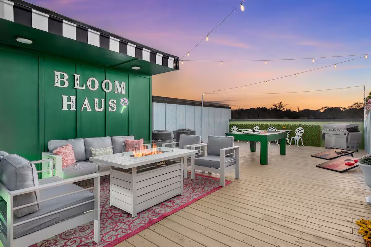 Rooftop deck at sunset with cozy outdoor seating around a white fire‑pit table, string lights overhead, green accent wall with floral sign, game table, cornhole boards and a barbecue grill.