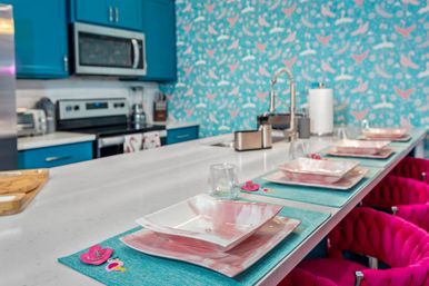 Bright, playful coastal-inspired kitchen with a white quartz island set with pink marbled square plates on turquoise placemats, teal cabinets, bird-pattern wallpaper, stainless appliances, and hot-pink bar stools.