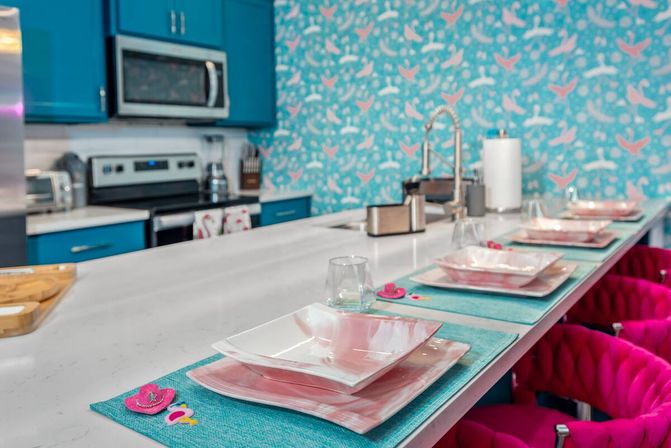 Bright, playful coastal-inspired kitchen with a white quartz island set with pink marbled square plates on turquoise placemats, teal cabinets, bird-pattern wallpaper, stainless appliances, and hot-pink bar stools.