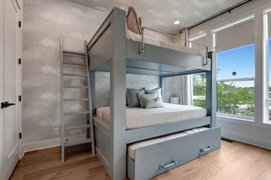 Cozy modern bedroom with a gray bunk bed and pull-out trundle, ladder, cloud-pattern wallpaper, light hardwood floors, and large windows showing green trees and blue sky.