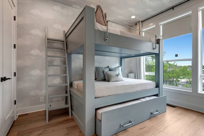 Cozy modern bedroom with a gray bunk bed and pull-out trundle, ladder, cloud-pattern wallpaper, light hardwood floors, and large windows showing green trees and blue sky.