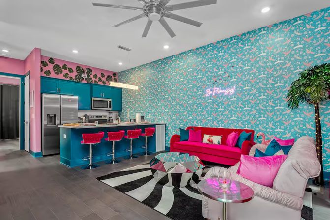 Playful tropical-themed open-plan living room and kitchen — teal cabinets, pink accents, hot-pink sofa, velvet bar stools, patterned teal wallpaper, neon wall sign, palm tree lamp and striped rug.