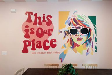 Playful pink dining-room wall mural with bold red lettering "This is Our Place — we make the rules" and a colorful pop-art portrait of a woman in sunglasses above a wooden table.