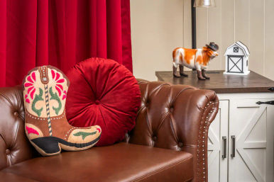Western home decor vignette: tufted brown leather sofa with decorative cowboy boot pillow and round red velvet cushion, white farmhouse cabinet topped with a cowboy-hat cow figurine and mini barn.