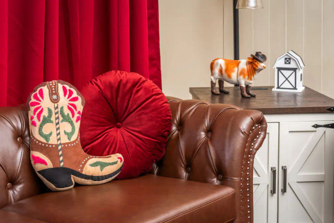 Western home decor vignette: tufted brown leather sofa with decorative cowboy boot pillow and round red velvet cushion, white farmhouse cabinet topped with a cowboy-hat cow figurine and mini barn.