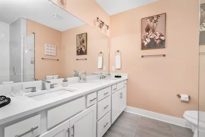 Contemporary peach bathroom with white double-sink vanity, quartz countertop, large mirror, glass shower, gray tile floor, and floral wall art.