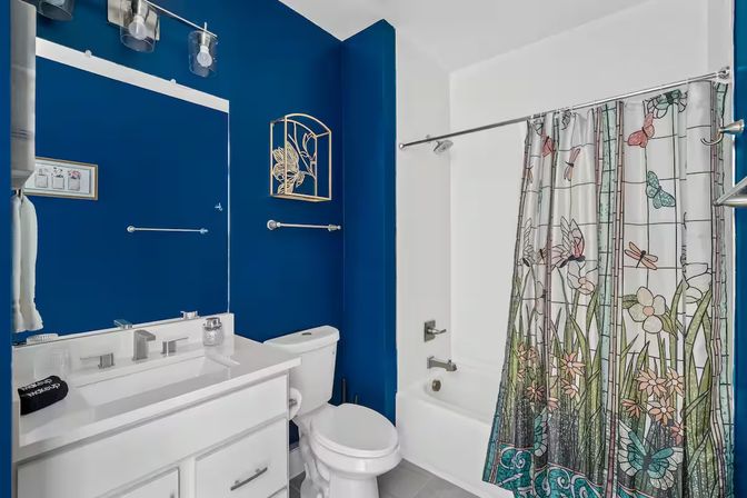 Modern bathroom with deep navy-blue walls, white vanity and toilet, chrome fixtures, and a tub with a stained-glass style floral and dragonfly shower curtain, accented by a gold wall decor and large mirror.