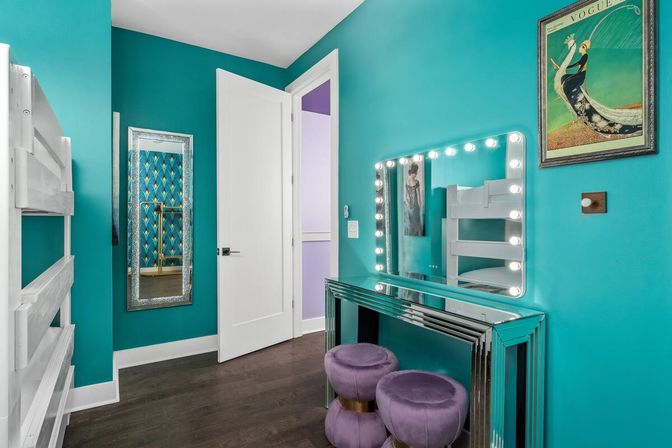 Vibrant turquoise bedroom featuring a white bunk bed, mirrored vanity with lit Hollywood-style mirror, two purple velvet poufs, vintage art print, decorative full-length mirror, and dark hardwood floor