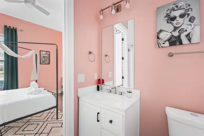 Cheerful powder-pink ensuite bathroom with a white modern vanity, chrome fixtures and mirror, retro black-and-white wall art, and an open doorway revealing a bedroom with a black canopy bed, dark green curtains and geometric rug.