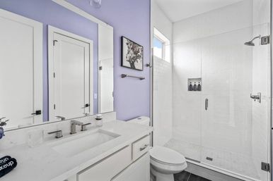 Bright modern home bathroom with lavender accent wall, white quartz vanity and undermount sink, large mirror, toilet, and frameless glass shower with white subway tile and chrome fixtures.