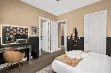 Modern bedroom interior with beige-and-black two-tone walls, hardwood floor, white bed with brown throw, illuminated vanity and record-themed wall art, tan accent chair, and open doorway to bathroom