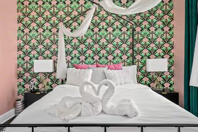 Art Deco–inspired bedroom with pink-and-green geometric wallpaper, white canopy bed with pink accent pillows, towel swans forming a heart on crisp white linens, black nightstands and modern lamps