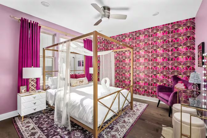 Vibrant pink and gold bedroom interior with a four‑poster canopy bed draped in sheer curtains, geometric magenta wallpaper, velvet armchair, gold‑accent nightstand and patterned area rug.